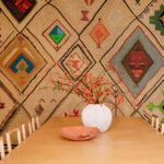 Eettafel met een witte vaas en oranje takken voor een wand bekleed met Elitis Hydra Lin Brodés VP 957 01 behang met kleurrijke geometrische patronen.