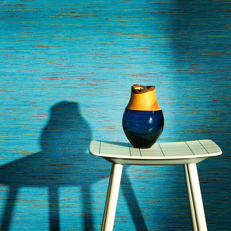 Blauw-geel keramieken vaas op een licht houten kruk tegen Elitis Indiana VP 851-1 behang in blauwe tinten.