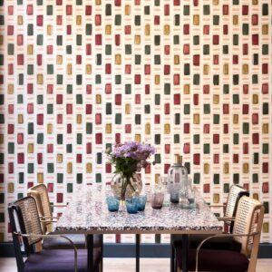 Eettafel met glazen decoraties en een vaas bloemen, tegen een kleurrijk Elitis Initiation Ornements-behang met rechthoekig patroon.