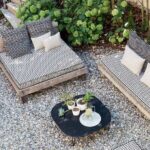 Twee loungebanken met Elitis Iseo outdoor stof en sierkussens staan op een grindterras naast een zwarte salontafel met planten en limonade.