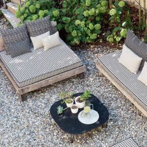Twee loungebanken met Elitis Iseo outdoor stof en sierkussens staan op een grindterras naast een zwarte salontafel met planten en limonade.