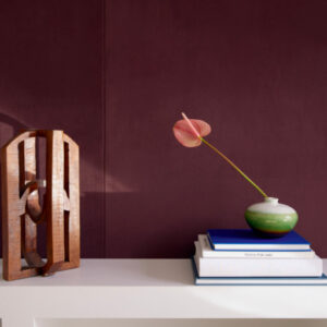 Decoratieve opstelling met een houten sculptuur, stapel boeken en vaas met bloem voor een donkerpaars Elitis Parthenon RM 1045 70 Rayures Jumelles behang.
