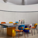 Moderne eetkamer met een betonnen tafel, kleurrijke stoelen en Elitis Rayures Jumelles behang op de achterwand.