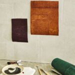 Collage met Elitis Sangpi VP 1018-60 behangstaal in warme tinten, omringd door schildermaterialen op een tafel.