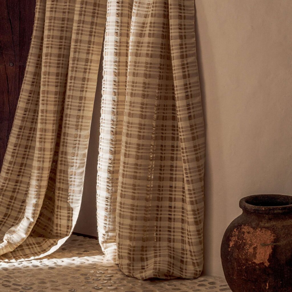 Lichtdoorlatend Elitis Baleares Brisa CN 004 29 gordijn met beige ruitpatroon in een rustieke interieursetting.