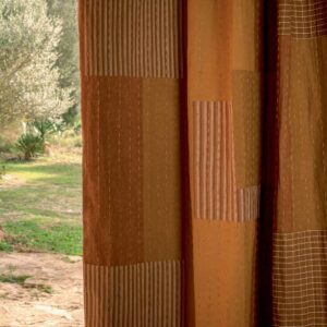 Oranje- en bruintinten Elitis Baleares Costa LZ 902 05 gordijn met textuur en patronen, hangend voor een tuinzicht.