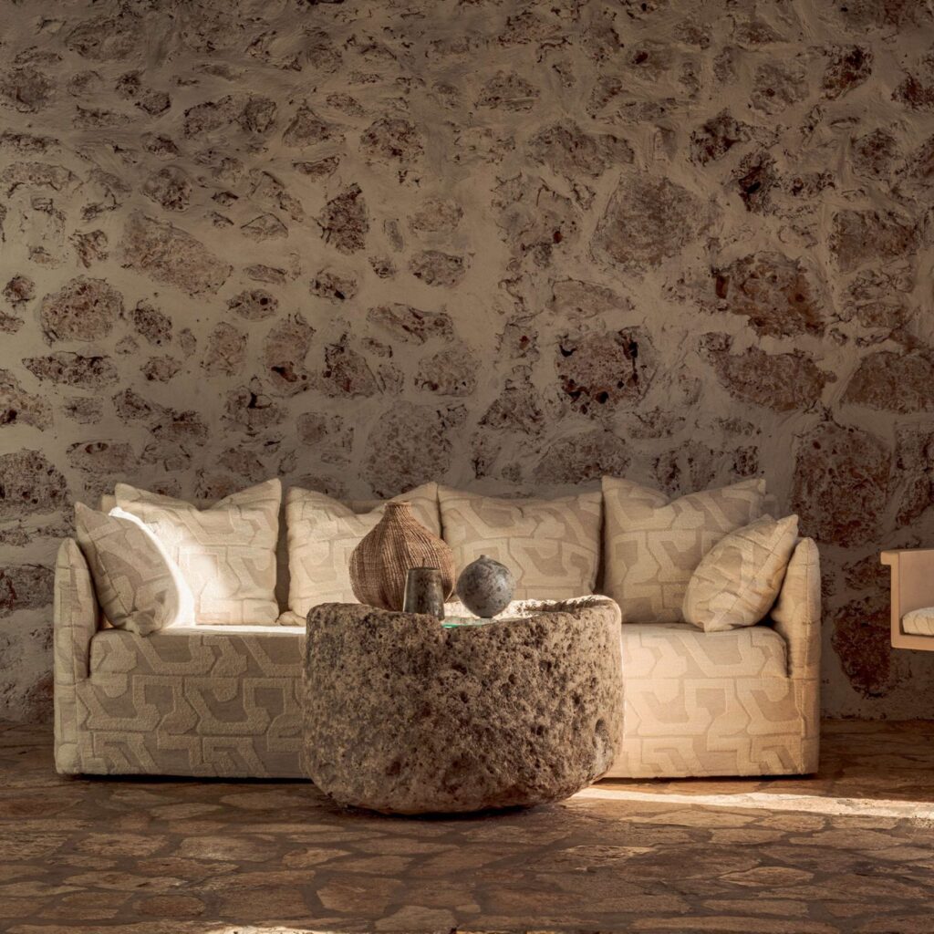 Beige Elitis Eivissa-bank in mediterrane sfeer met een robuuste, ronde stenen salontafel tegen een rustieke stenen muur.