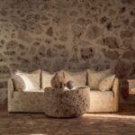 Beige Elitis Eivissa-bank in mediterrane sfeer met een robuuste, ronde stenen salontafel tegen een rustieke stenen muur.