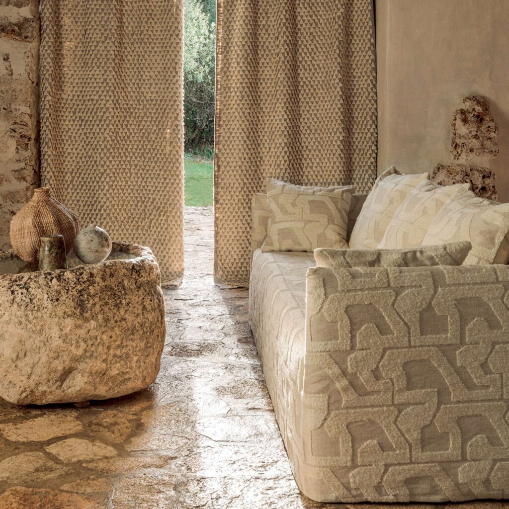 Beige woonkamer met Elitis Baleares Eivissa LR 370 01 stoffen op de bank en gordijnen, met een rustieke stenen tafel op een natuurstenen vloer.