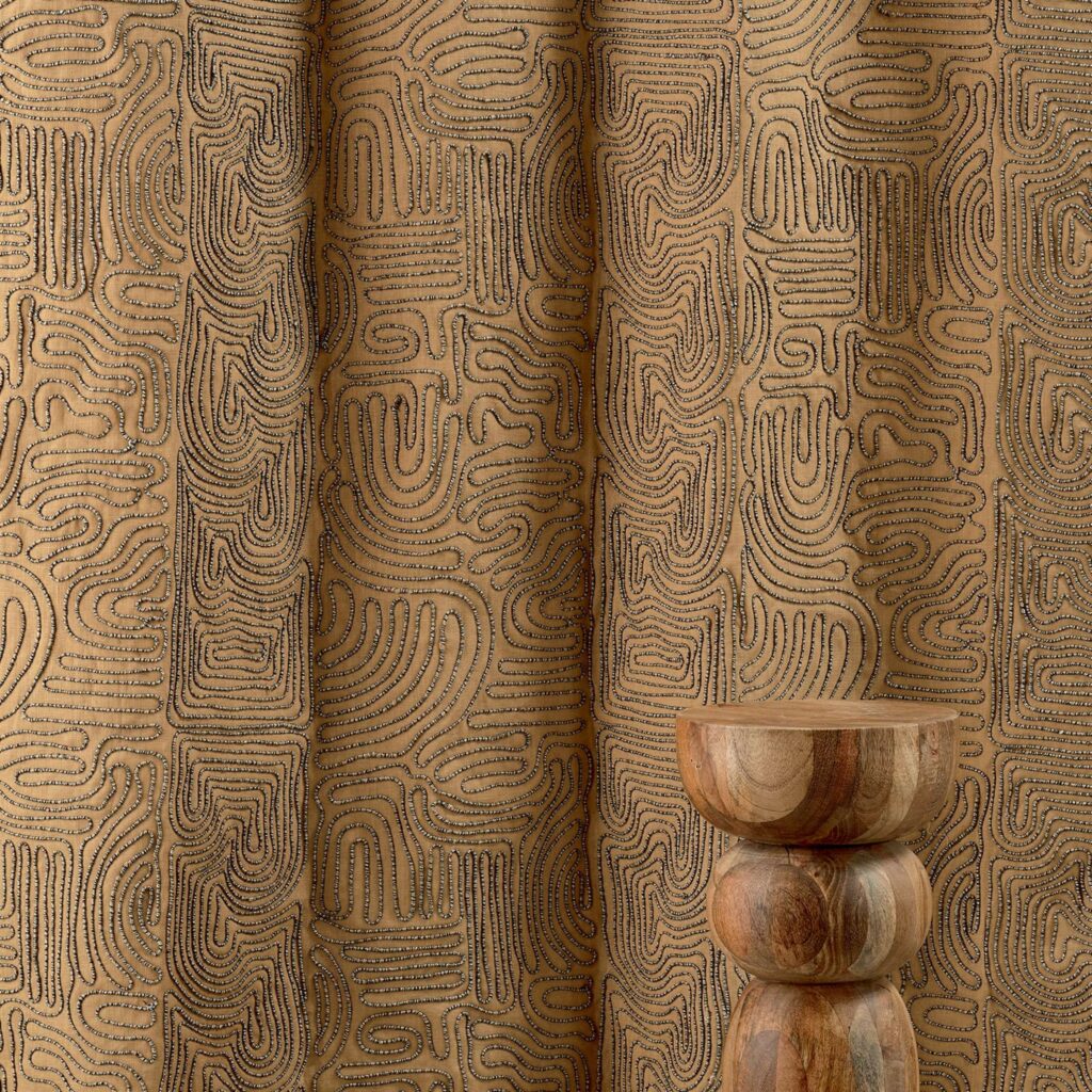 Beige Elitis Baleares Llenya LZ 905 01 gordijn met abstract patroon en houten decoratief object op de voorgrond.