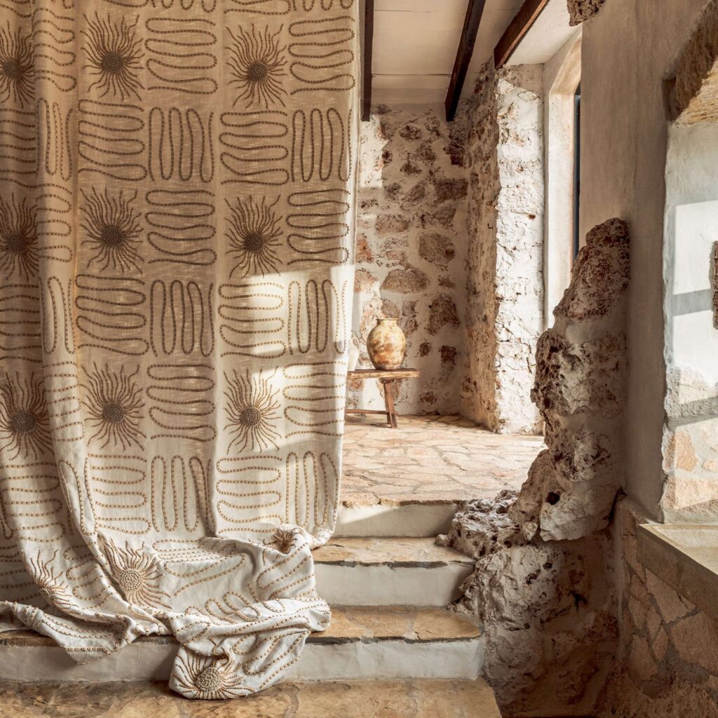 Gordijn uit de Elitis Baleares Paloma LZ 898 01 collectie in een rustieke ruimte met natuurstenen muren en een aardewerken vaas op een houten krukje.