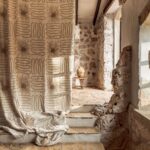 Gordijn uit de Elitis Baleares Paloma LZ 898 01 collectie in een rustieke ruimte met natuurstenen muren en een aardewerken vaas op een houten krukje.