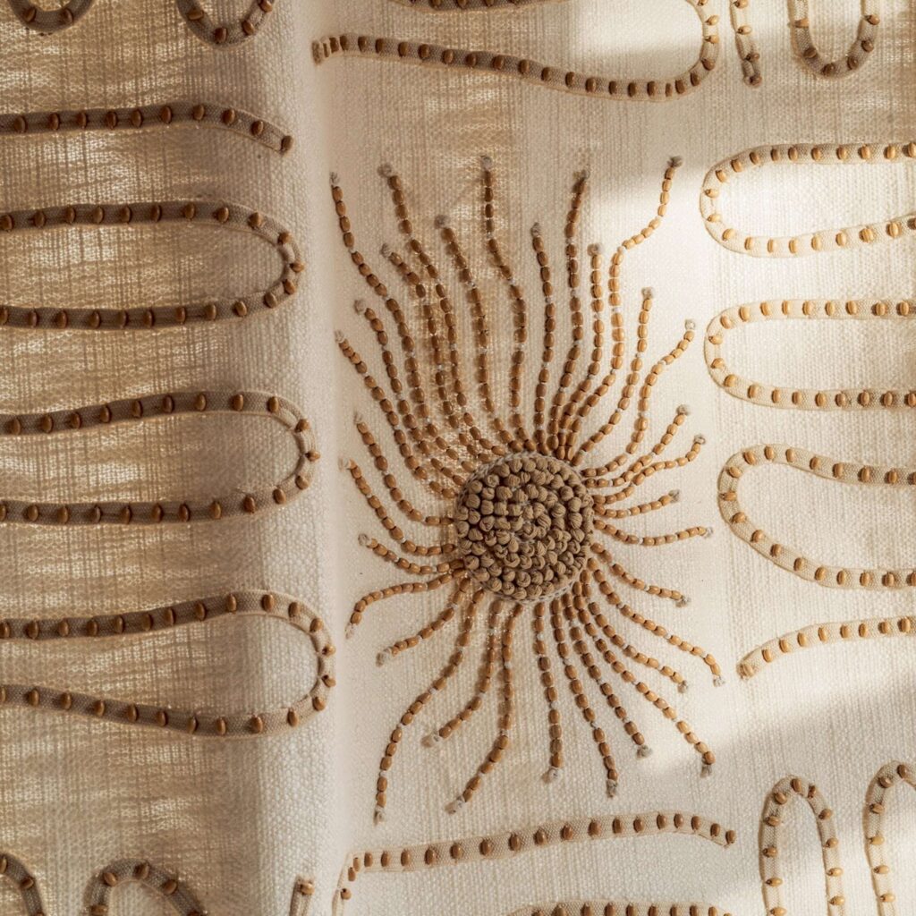 Detail van Elitis Baleares Paloma LZ 898 01 stof met beige geborduurd zonmotief en golvende patronen op een lichtdoorlatende ondergrond.