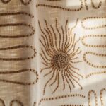 Detail van Elitis Baleares Paloma LZ 898 01 stof met beige geborduurd zonmotief en golvende patronen op een lichtdoorlatende ondergrond.