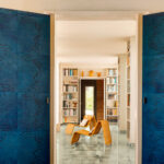 Moderne woonkamer met blauw Elitis Art Paper Jima behang en rotan loungestoelen tussen open boekenkasten.