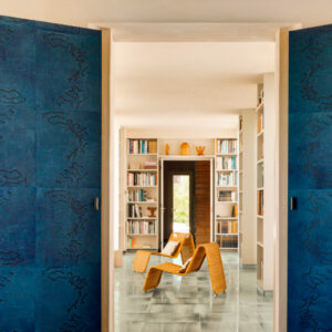 Moderne woonkamer met blauw Elitis Art Paper Jima behang en rotan loungestoelen tussen open boekenkasten.