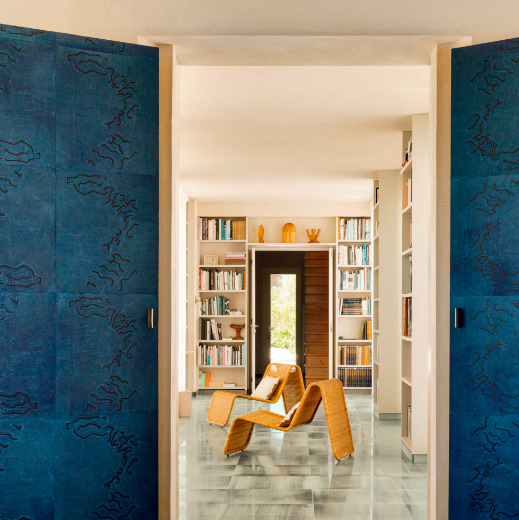 Moderne woonkamer met blauw Elitis Art Paper Jima behang en rotan loungestoelen tussen open boekenkasten.