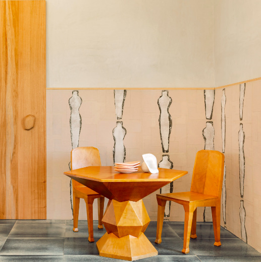 Houten tafel en stoelen in een kamer met Elitis behang ‘Naoshima RM 1031 03’ met abstracte figuren op de muur.