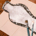 Abstracte vorm op Elitis-behang Art Paper Naoshima RM 1031 03 met twee verfkwasten erop geplaatst.