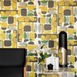 Kleurrijk Elitis-behang Chaman TP315 02 met geometrisch patroon in geel, groen, grijs en wit achter een modern interieur met zwarte stoel en decoratieve accessoires.