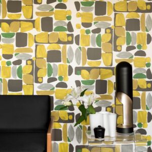 Kleurrijk Elitis-behang Chaman TP315 02 met geometrisch patroon in geel, groen, grijs en wit achter een modern interieur met zwarte stoel en decoratieve accessoires.