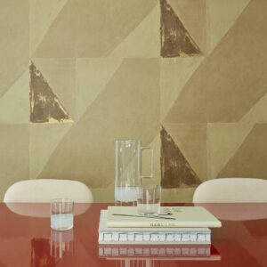 Interieur met Elitis-behang uit de Voile de Papier collectie, geometrisch patroon in aardetinten, met glazen karaf, drinkglazen en boeken op een glanzende rode tafel.