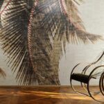 Behang uit de Elitis Cuba Libre collectie met een groot palmbladmotief en een rotan schommelstoel op een houten vloer.