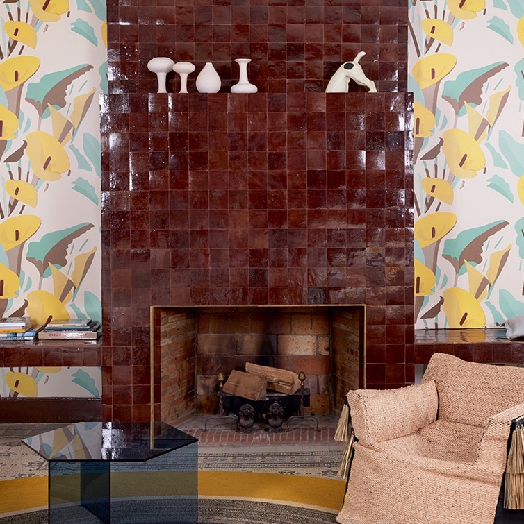 Woonkamer met een roodbruine haard, Elitis-behang Flower Power Arum en een rotan fauteuil.