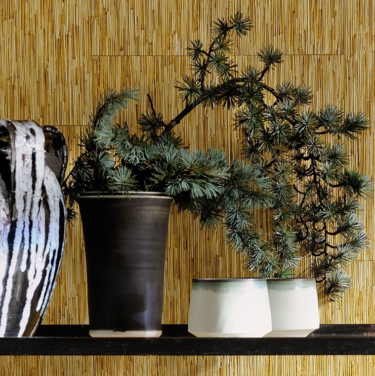 Keramieken vazen en bekers met dennentakken staan tegen een muur met Elitis Formentera strobehang.