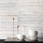 Zachte witte muur met Elitis-behang Iloilo RM 970 01 als achtergrond, met witte kommen en gouden kandelaars op een donker meubel.