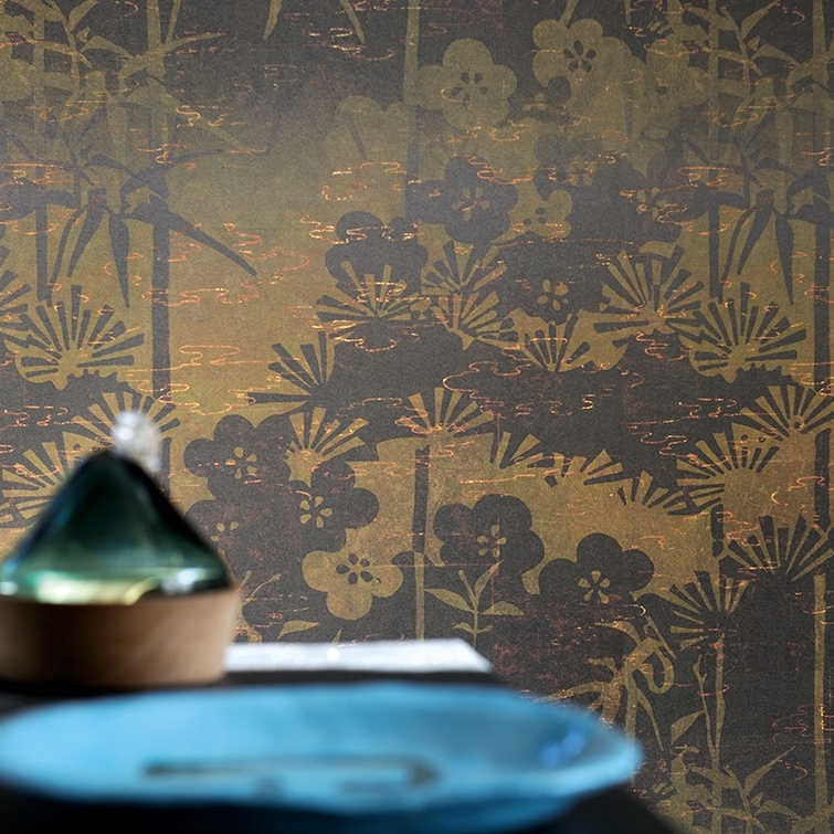 Elitis-behang La Montagne Sacrée met een botanisch patroon in goud- en bruintinten op de achtergrond van een gedekte tafel.