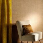 Luxe Elitis-behang uit de Madagascar-collectie met een beige textuur, naast een lichtgrijze stoel en gele gordijnen.