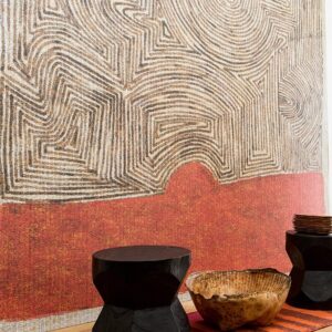 Elitis-behang Mexicas VP917-01 met abstract patroon in aardetinten tegen een wand met houten meubels op de voorgrond.