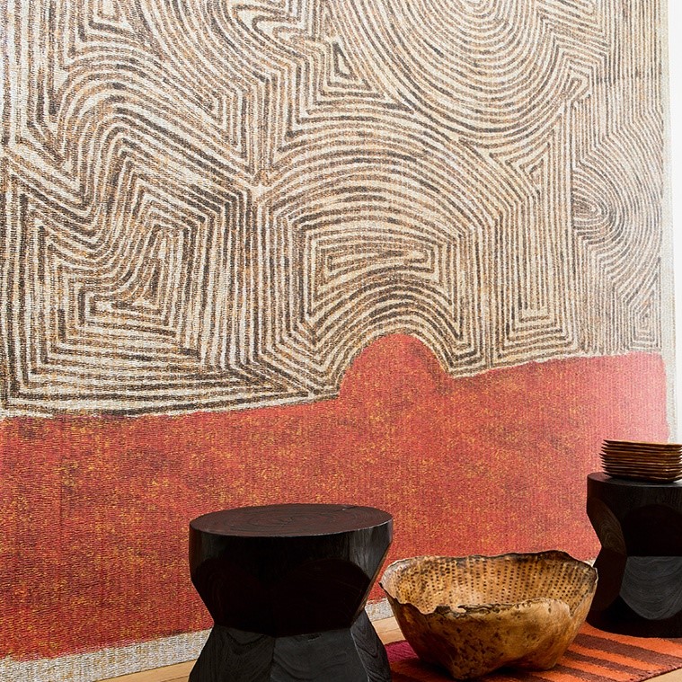 Elitis-behang Mexicas VP917-01 met abstract patroon in aardetinten tegen een wand met houten meubels op de voorgrond.