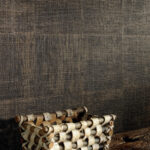 Donker Elitis-behang Raffia VP 601 80 met grove textuur, gecombineerd met een decoratief houten mandje op de voorgrond.