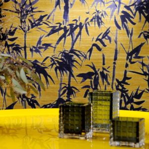 Gele wand met Elitis-behang Talamone Terra Promessa VP 854 02 met donkerblauw bamboepatroon en glazen decoratie op een gele tafel.
