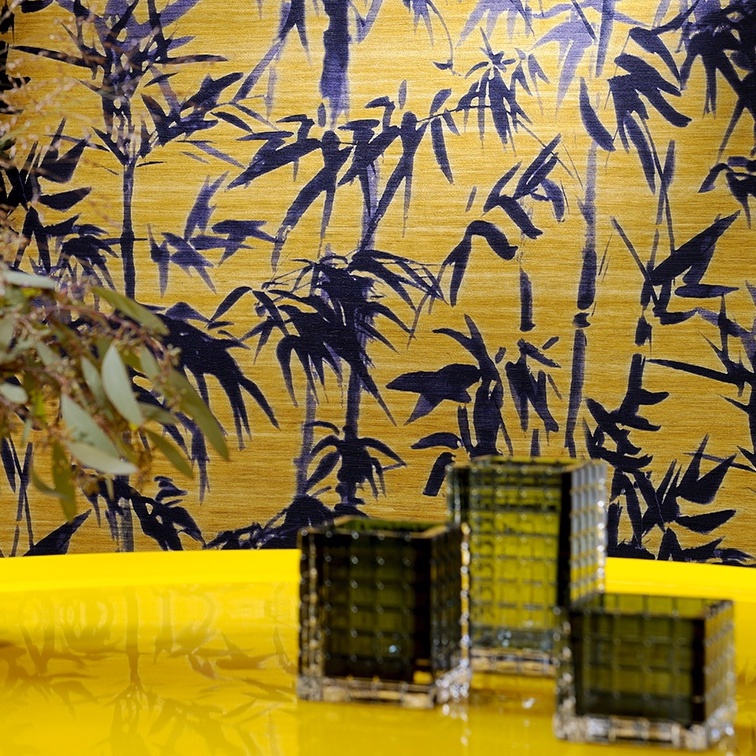 Gele wand met Elitis-behang Talamone Terra Promessa VP 854 02 met donkerblauw bamboepatroon en glazen decoratie op een gele tafel.