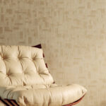 Beige Elitis-behang Bois Sculpte Ramille VP 938 20 met abstract reliëfpatroon achter een crèmekleurige, gecapitonneerde designstoel.