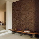 Moderne interieurhoek met Elitis-behang Bois Sculpté Ramille VP 938 71 en een houten designbank tegen een donkerbruin geometrisch patroon.