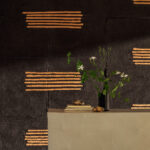 Zwarte muur met Elitis Hanji Noto RM 1058 80 behang en horizontale koperkleurige accenten, met een vaas bloemen en boeken op een beige console.