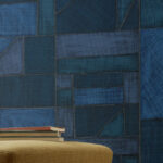 Geometrisch Elitis-behang La Caravane Kungbo VP 944 in verschillende blauwtinten met een poef en stapel boeken op de voorgrond.