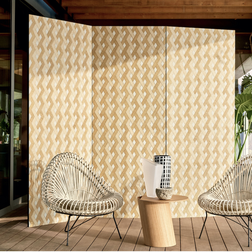 Twee rotan stoelen en een houten bijzettafeltje met vazen staan voor Elitis Merida Wall Entrelazados RM 1019 behang met een gevlochten patroon.
