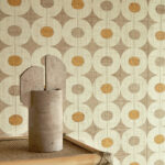 Decoratief Elitis behang uit de Merida Wall Pepite RM 1018 collectie met geometrisch patroon in aardetinten en oranje accenten, met een modern kunstobject op een rotan meubel op de voorgrond.