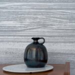 Donker keramieken vaasje op een witte schaal voor Elitis Mouvements Esprit Libre behang met grijs-wit houtnerfpatroon.