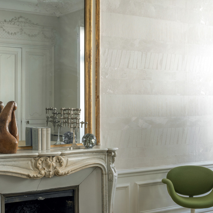Luxe interieur met Elitis-behang Mouvements Impulsion RM 1023-80 in neutrale tinten, een marmeren schouw en een groene designstoel.