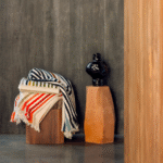 Gestreepte Elitis Betty PL 308 deken ligt over een houten kruk naast een zwarte vaas op een terracotta sokkel, tegen een donkere houten muur.