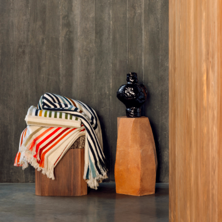 Gestreepte Elitis Betty PL 308 deken ligt over een houten kruk naast een zwarte vaas op een terracotta sokkel, tegen een donkere houten muur.