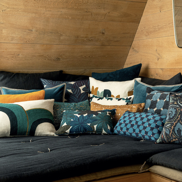 Sierkussens in diverse kleuren en patronen, waaronder het Elitis Bridge CO 216 46 02 kussen, op een donker bankje tegen een houten wand.