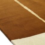 Detail van het Elitis Celeste TA 133 25 02 vloerkleed met een warme bruine kleur, een lichte middenstreep en een wit hoekvlak.
