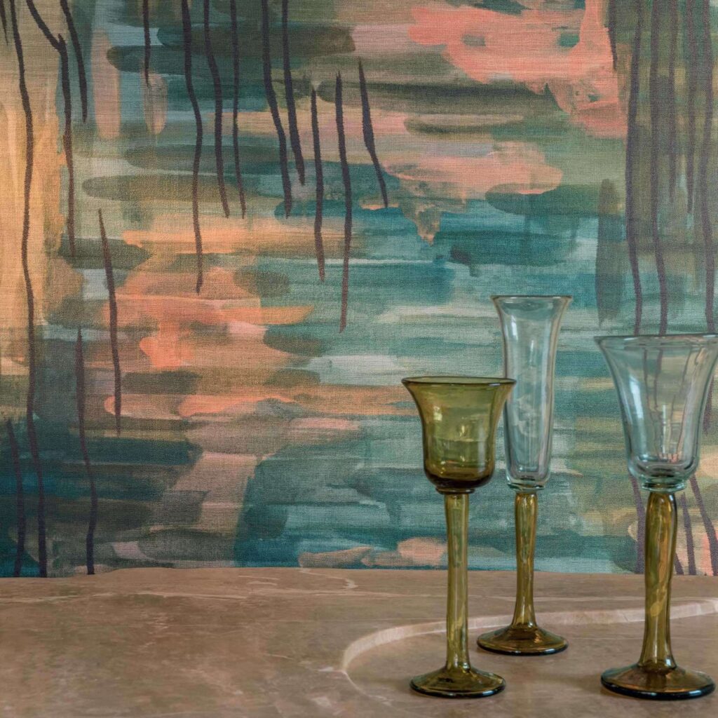 Detail van het Elitis Ceres Brocéliande behang met abstract geschilderd patroon in groene en roze tinten, met drie hoge glazen op een marmeren oppervlak.